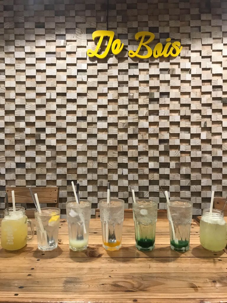 Seven glass of De Bois beverages in one line on the table with De Bois wooden bricks wall as background
